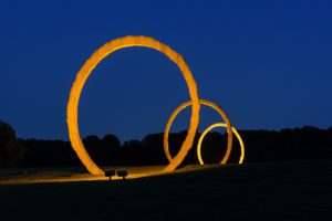 Three monumental, terracotta rings are lit orange at night against a midnight blue sky.