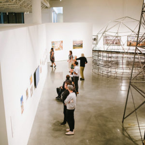 Visitors in a white-walled gallery at SECCA look at photographs and metal rod sculpture by Navajo artist Will Wilson.