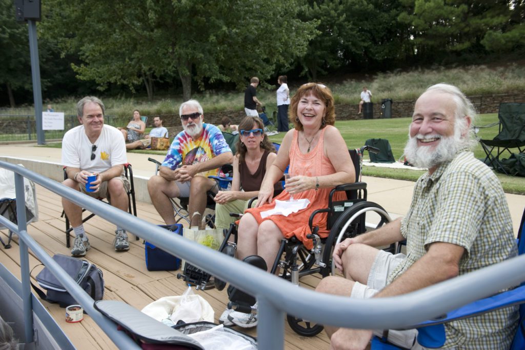 A group of smiling people sit in chairs and wheelchairs in the Museumâ€™s outdoor amphitheater accessible seating area.