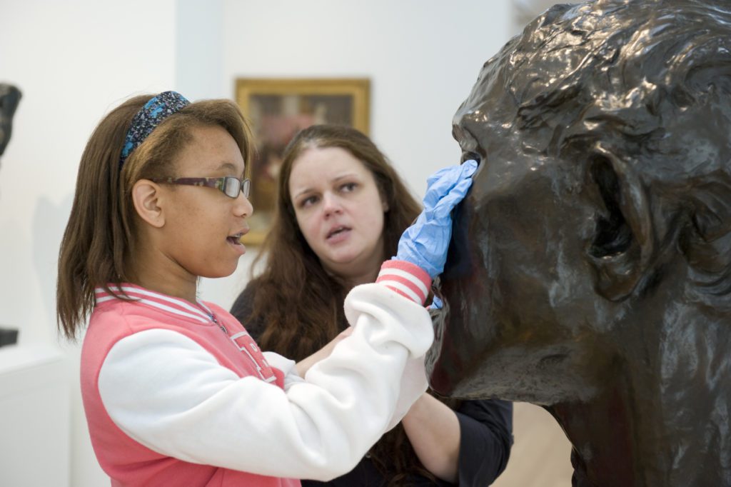 Accompanied by a tour guide, a student with a visual impairment wears gloves and touches a sculpture by French artist Auguste Rodin.