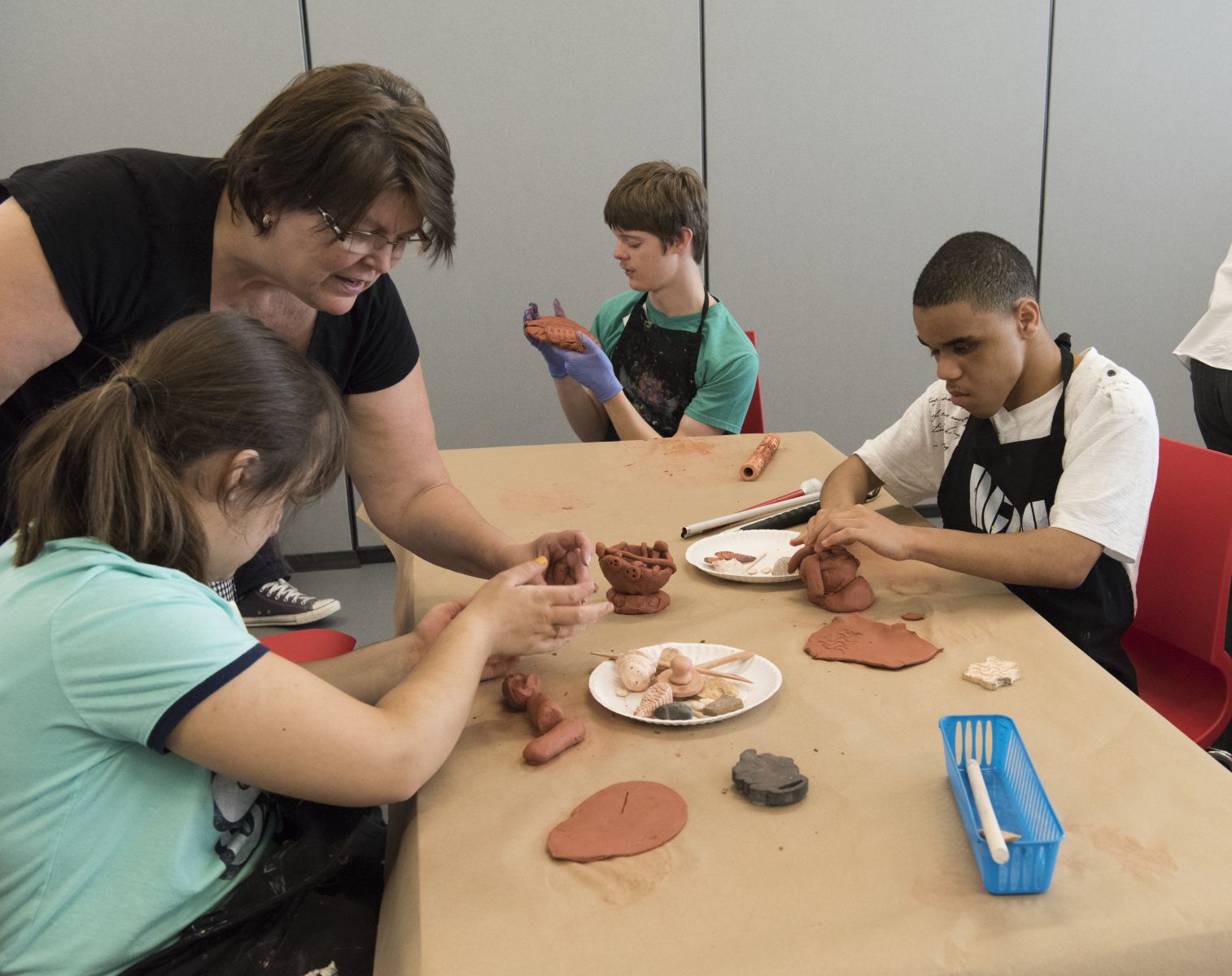 Three students sit at a table and work with clay with the help of a teacher.