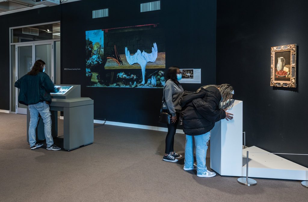 Three museum visitors are interacting with different sensory stations. A person on the left is drawing on a screen. Two people on the right are bent down leaning over a podium. 
