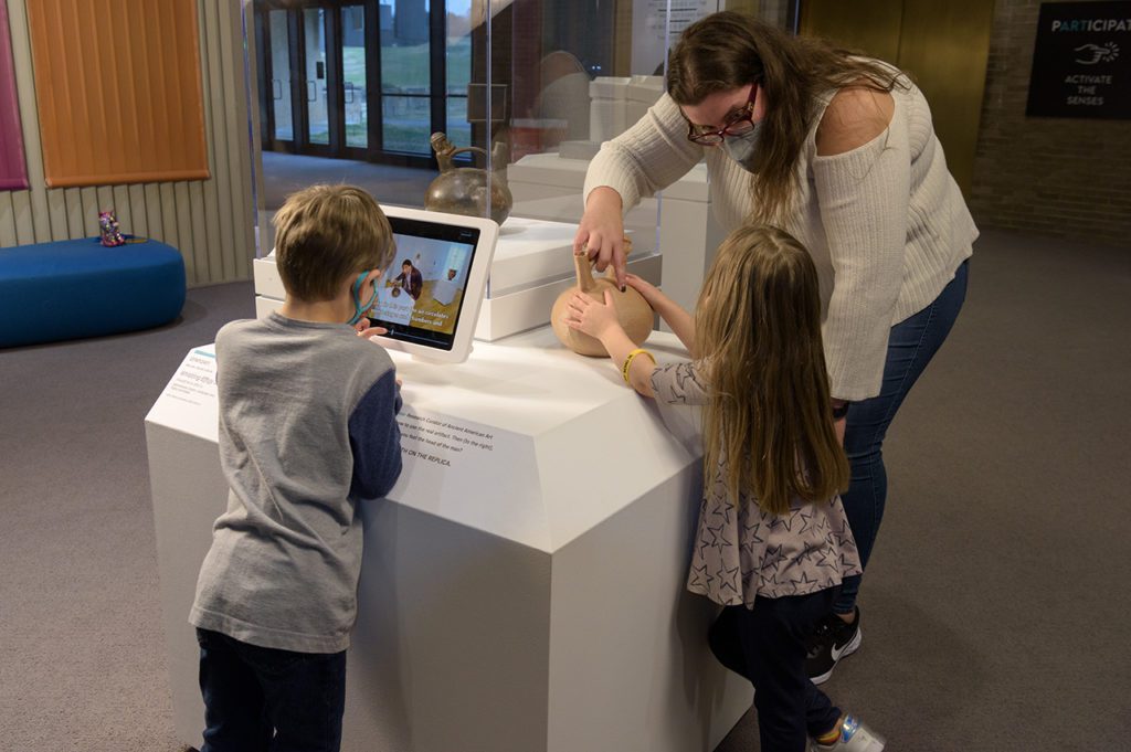 Two young children stand in front of an art station. One is watching a video, the other touches a replica of an artwork while talking to an adult on her right. 