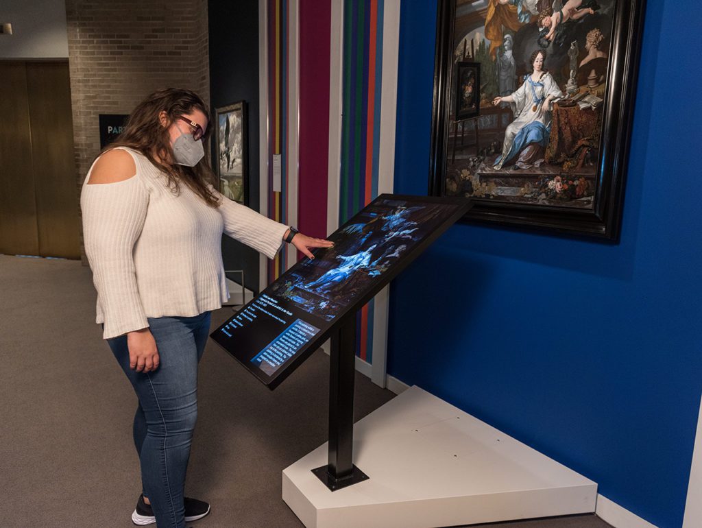 A women looks down and reaches out to touch a monitor with a replica of a painting attached to it. On the wall in front of her hangs the same painting. 