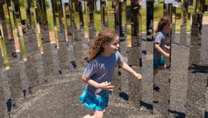 Young girl in a blue skirt dashes between mirrored pillars of a labyrinth sculpture.