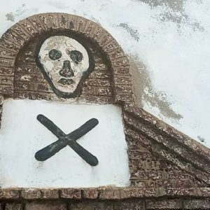Photo by artist Paa Joe of a mantel over a door at Elmina Castle in Ghana; a white skull with black eyes, nose, and mouth floats over a white square with a black X in the middle.