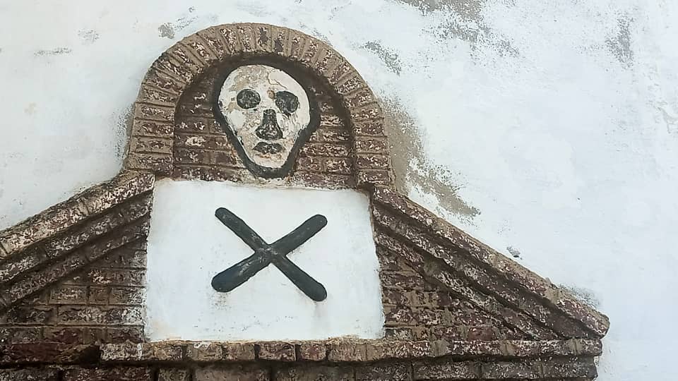 Photo by artist Paa Joe of a mantel over a door at Elmina Castle in Ghana; a white skull with black eyes, nose, and mouth floats over a white square with a black X in the middle.