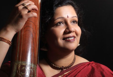 Bust-length photographic portrait of a woman in a red sari holding the neck of a stringed instrument.