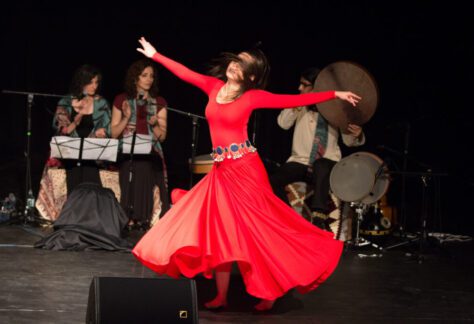 A woman in a long red dress twirls around with her arms out while two woman clap and a percussionist plays in the background.