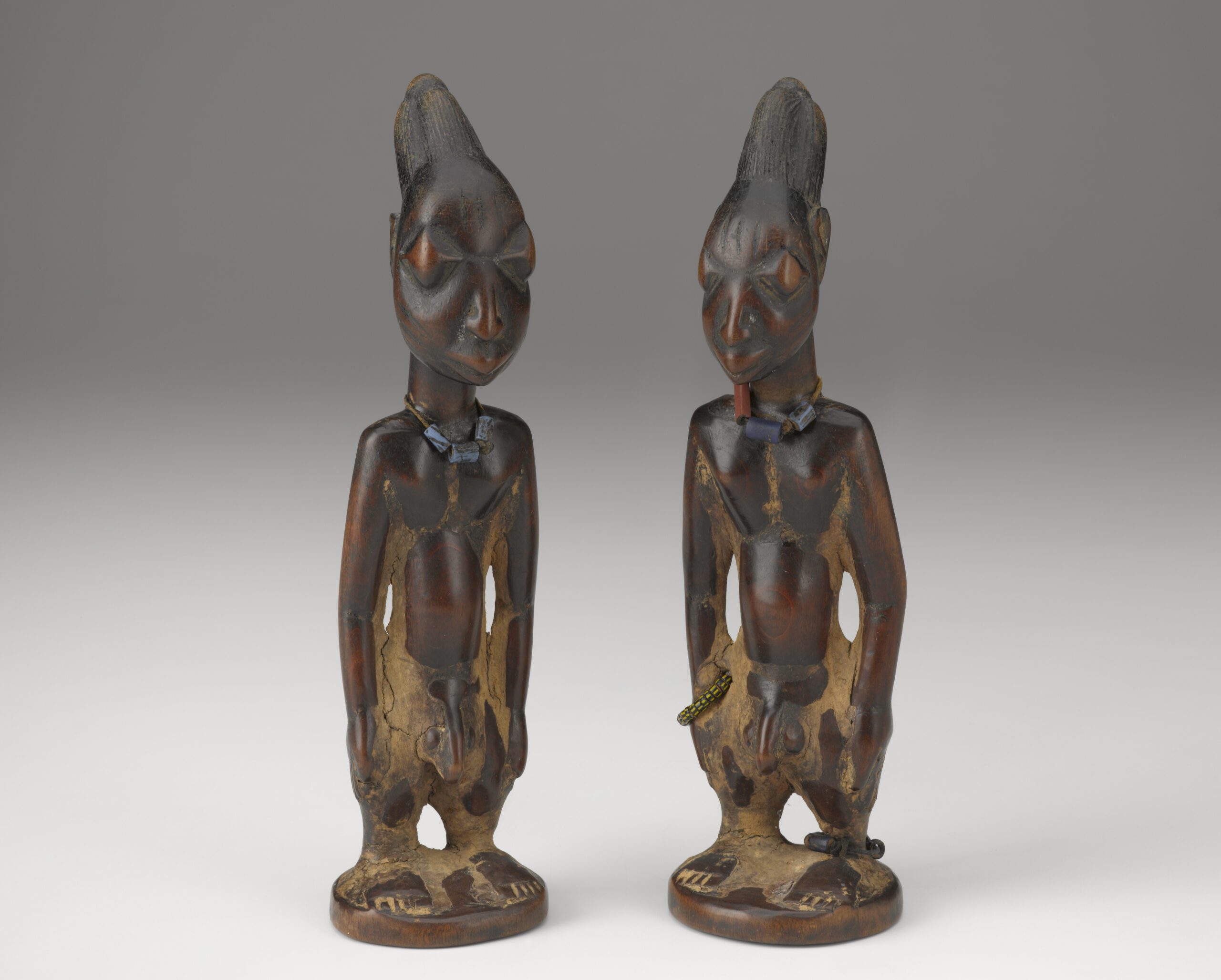 Two nearly identical wooden figures with large heads, elongated hairstyles, erect postures, and large eyes. 