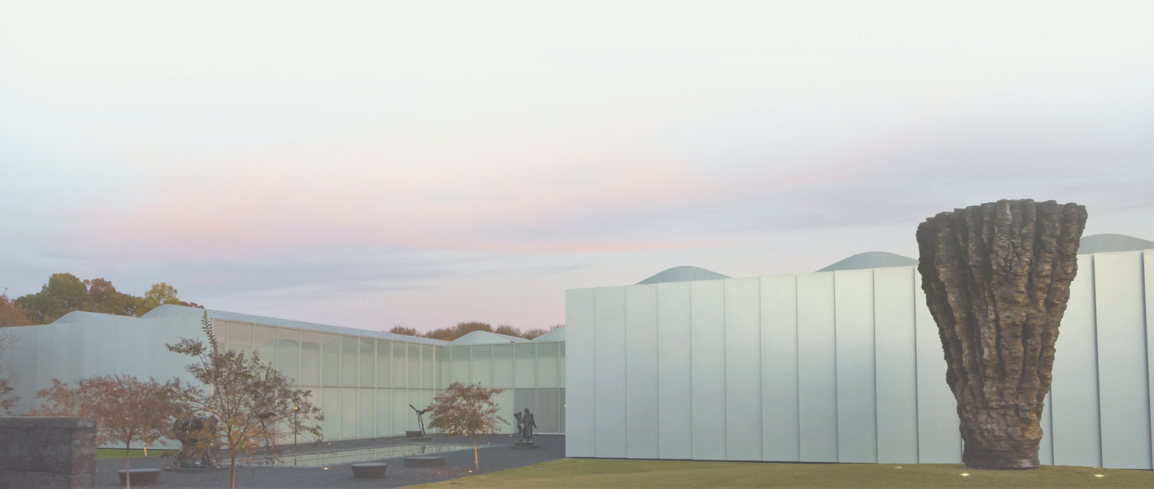 A tall, craggy, abstract sculpture in front of a sprawling aluminum and glass building with a courtyard.