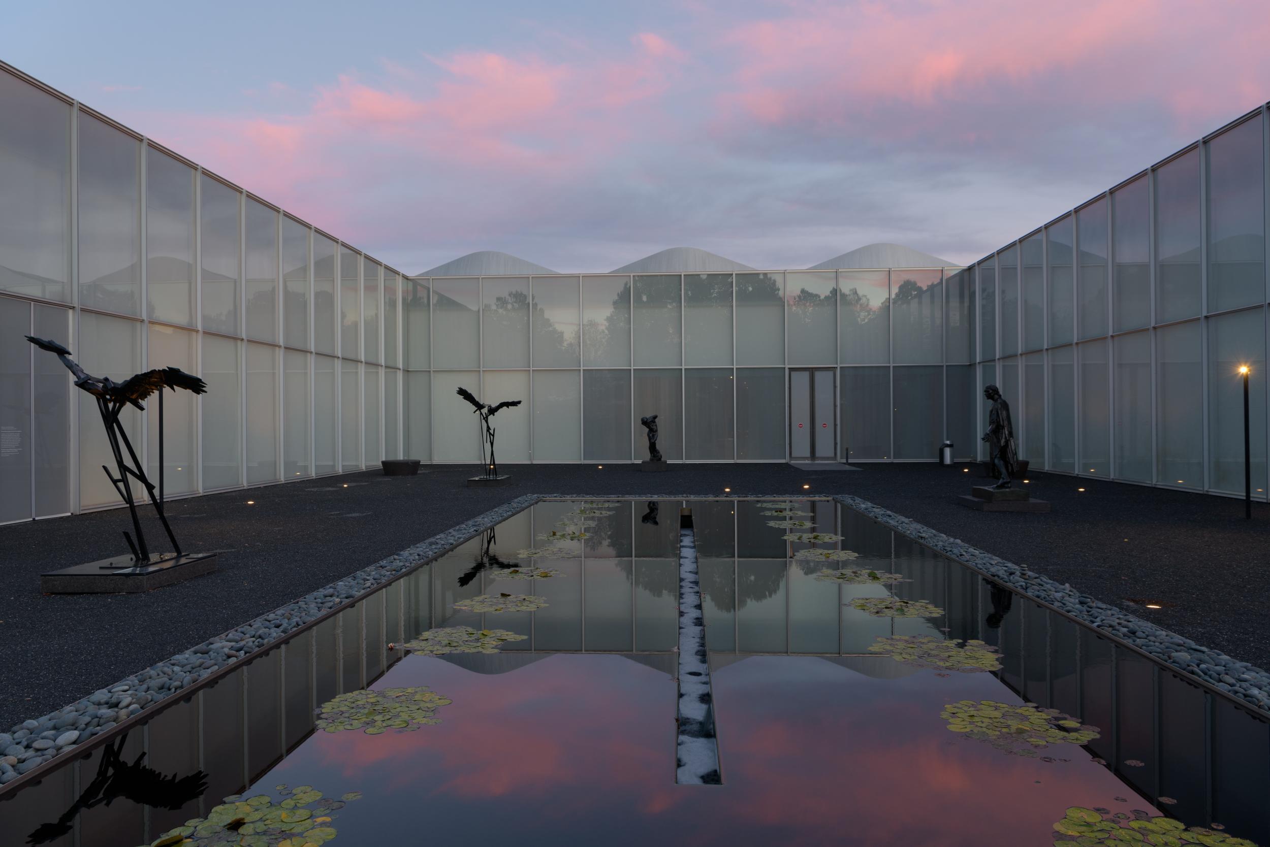 An outdoor courtyard at dusk with bronze sculptures organized around a large reflecting pool.