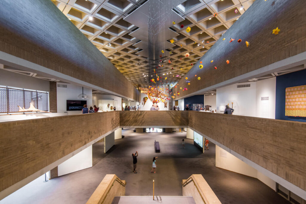 The interior atrium of a three-story building, decorated with art of various media and flowers suspended from the ceiling.