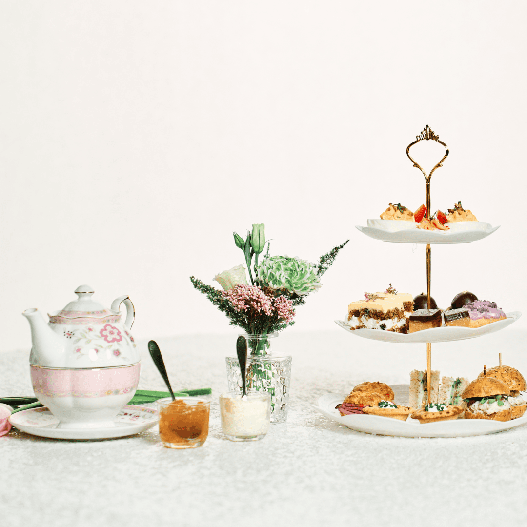 A ceramic teapot, flower arrangement, and tiered platter of sandwiches and desserts sits on a white tablecloth.