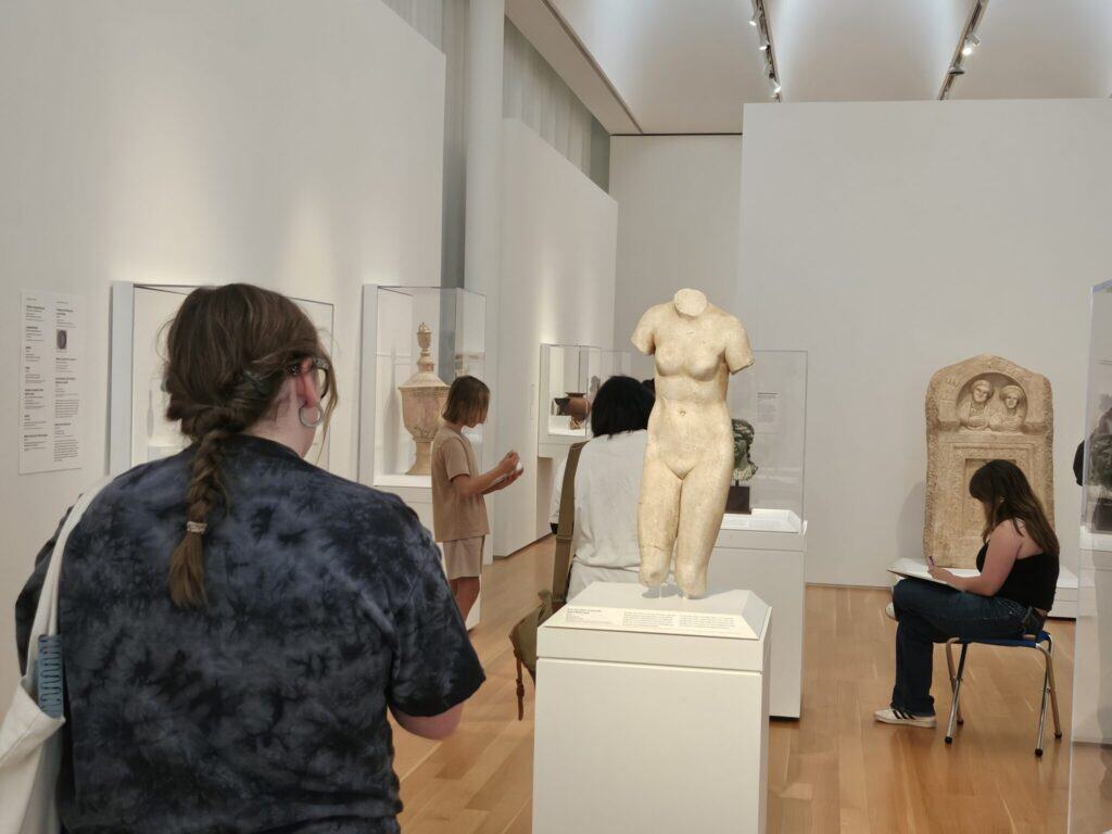 A back view of a teen sketching a nude marble sculpture of a woman's torso and upper legs. In the background other teens sketch a partially obstructed work of art.