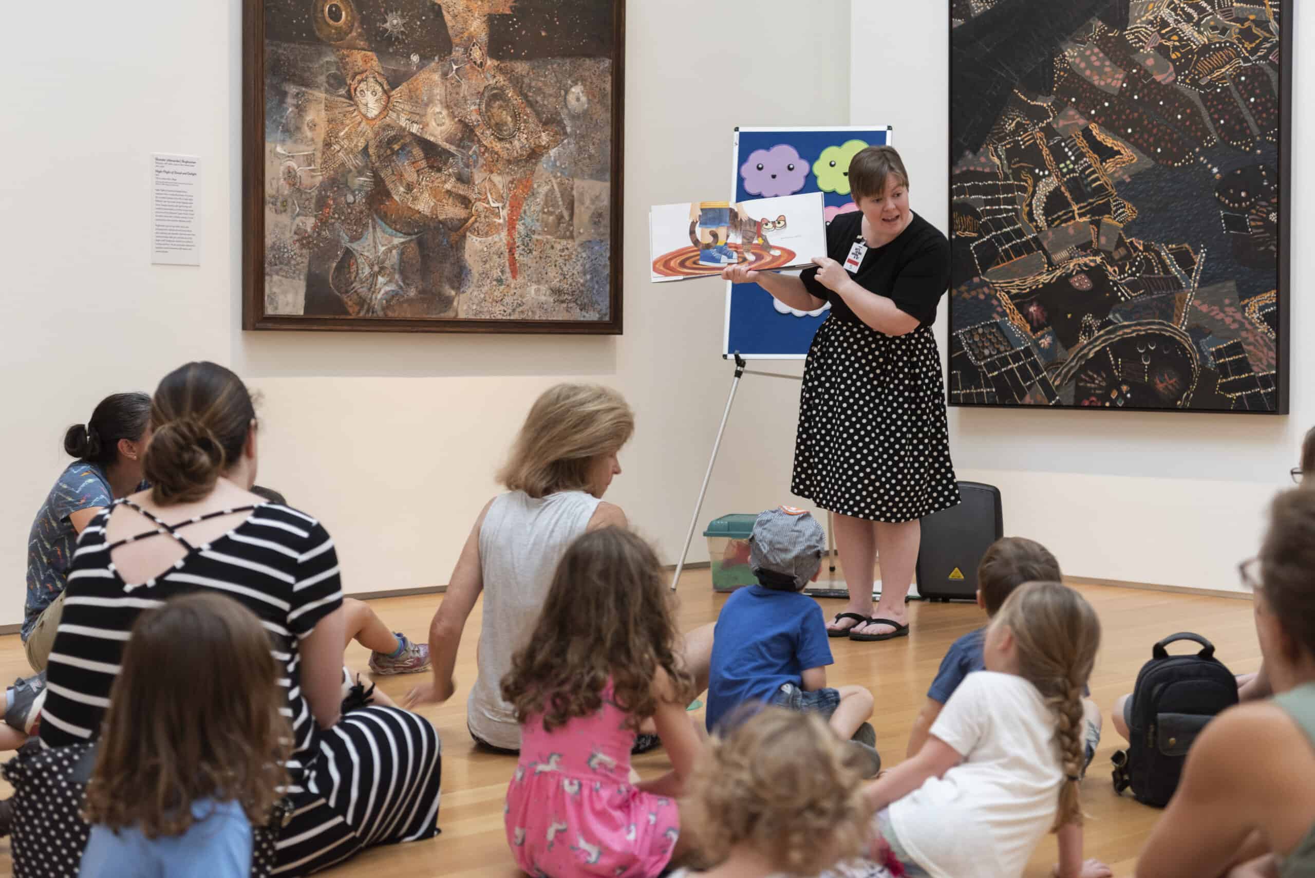 Adults and children sit on the floor of an art gallery and listen to a story reader showing a children’s book.