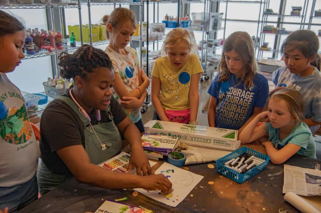 A group of children gather around a person wearing an apron and drawing with pastels on a piece of white paper. 