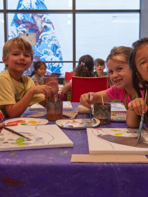 Four children wearing smocks pose while painting canvases on a table with a purple tablecloth. In the background there is a white brick wall decorated with a blue and purple oceanic mural.