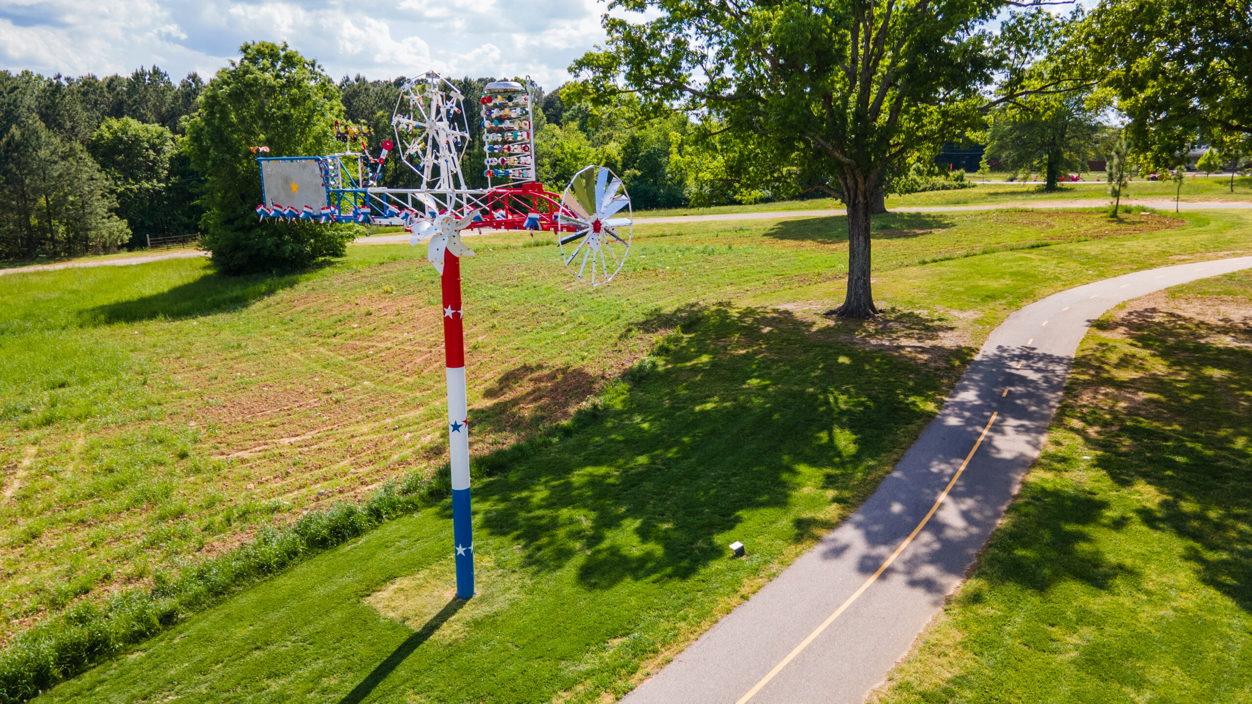 A red, white, and blue, composite sculpture resembling a whirligig installed along a paved park path.