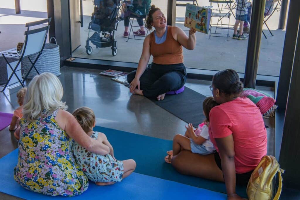 Two people sitting on the ground with children on their laps face someone sitting cross-legged and displaying an illustrated book. 