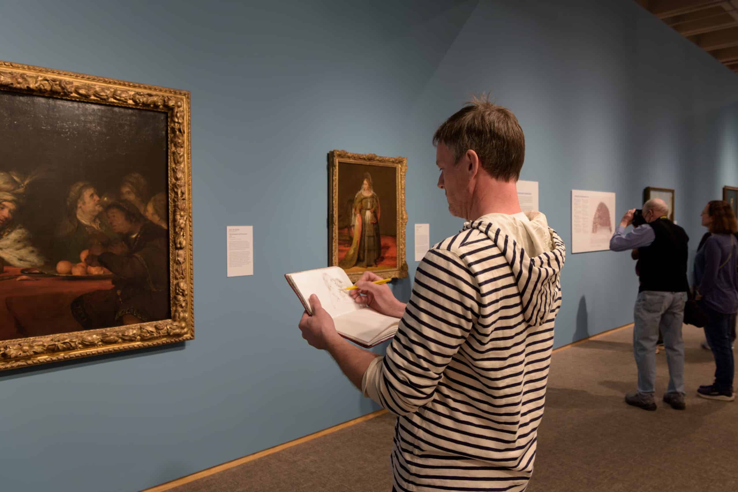A man wearing a stripped hoodie sketches a painting in an art gallery. 