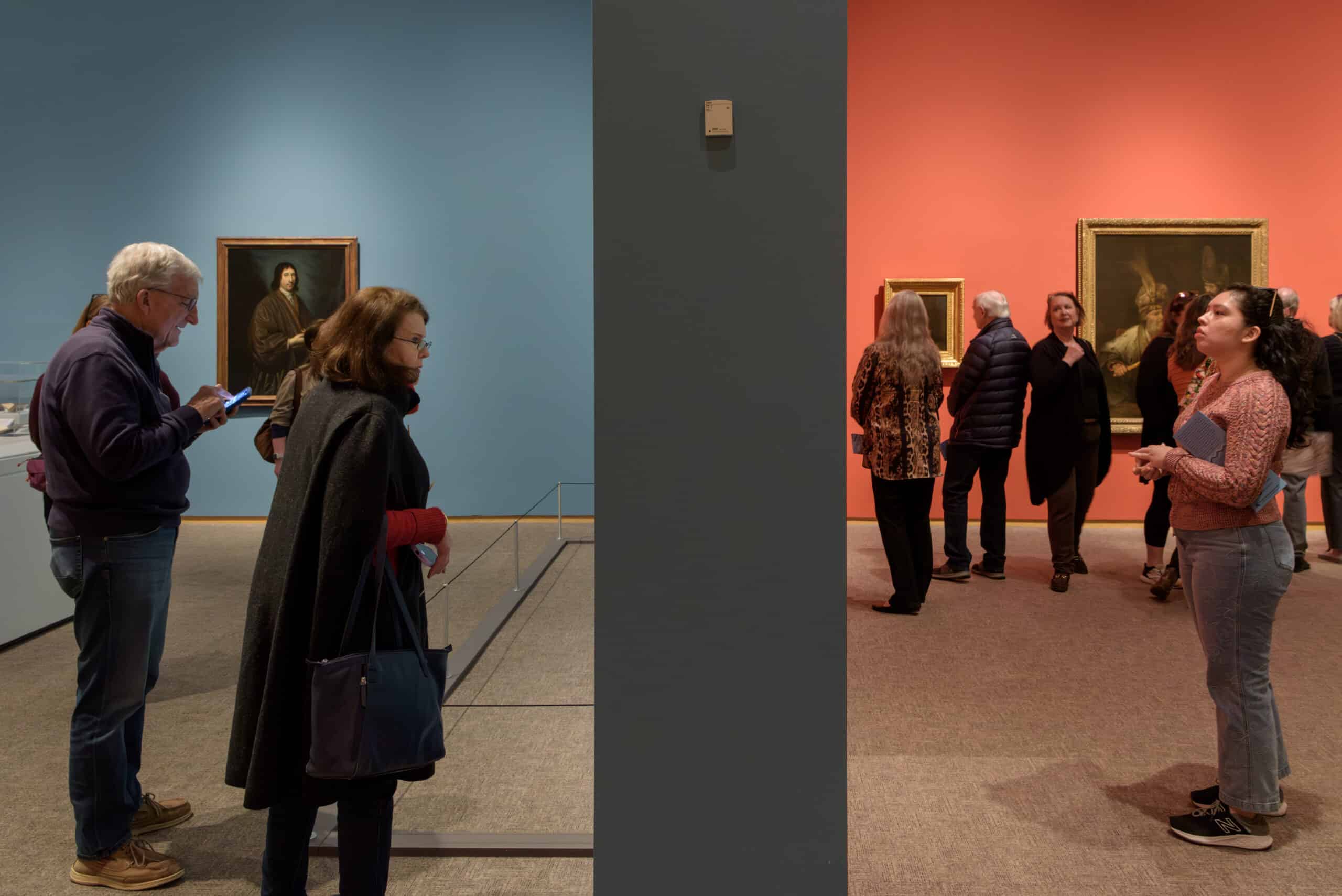 Two rooms in an art gallery, one blue and one red, are separated by a gray wall. In both of the two rooms, people observe and enjoy artwork. 