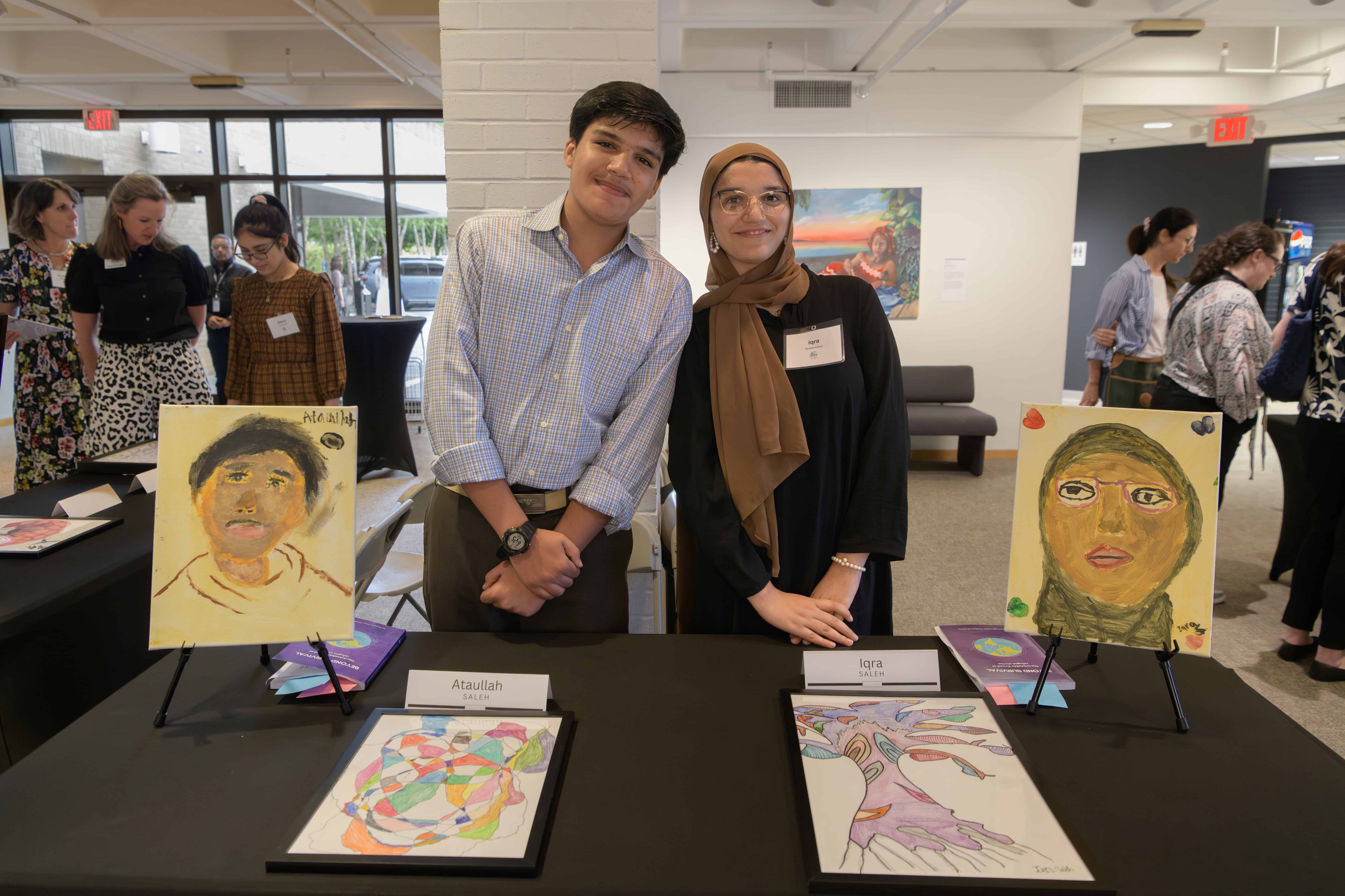 Two people pose behind a black table displaying illustrated artwork and painted portraits. 