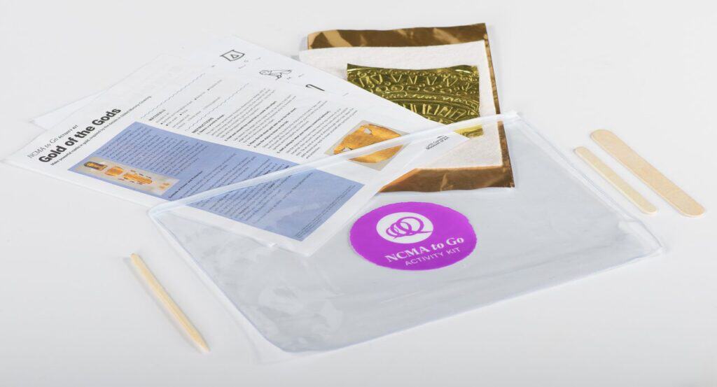 A clear plastic folder with papers sticking out of the top. A sticker on the folder reads, "NCMA to Go Art Activity Kit." Craft sticks lie on either side of the folder. 