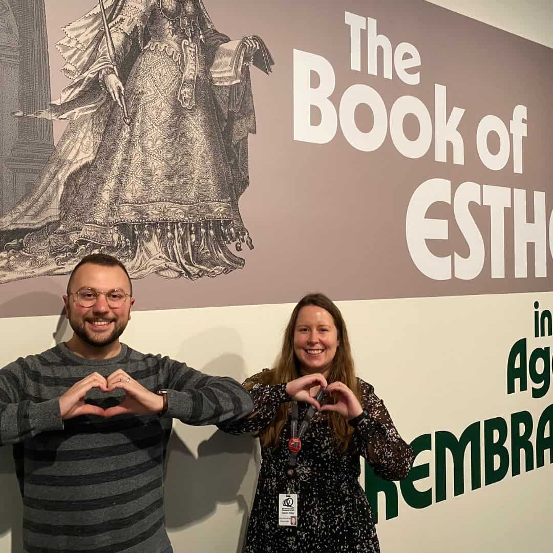 Two people making the shape of a heart with their hands stand in front of a beige and gray wall with writing on it.