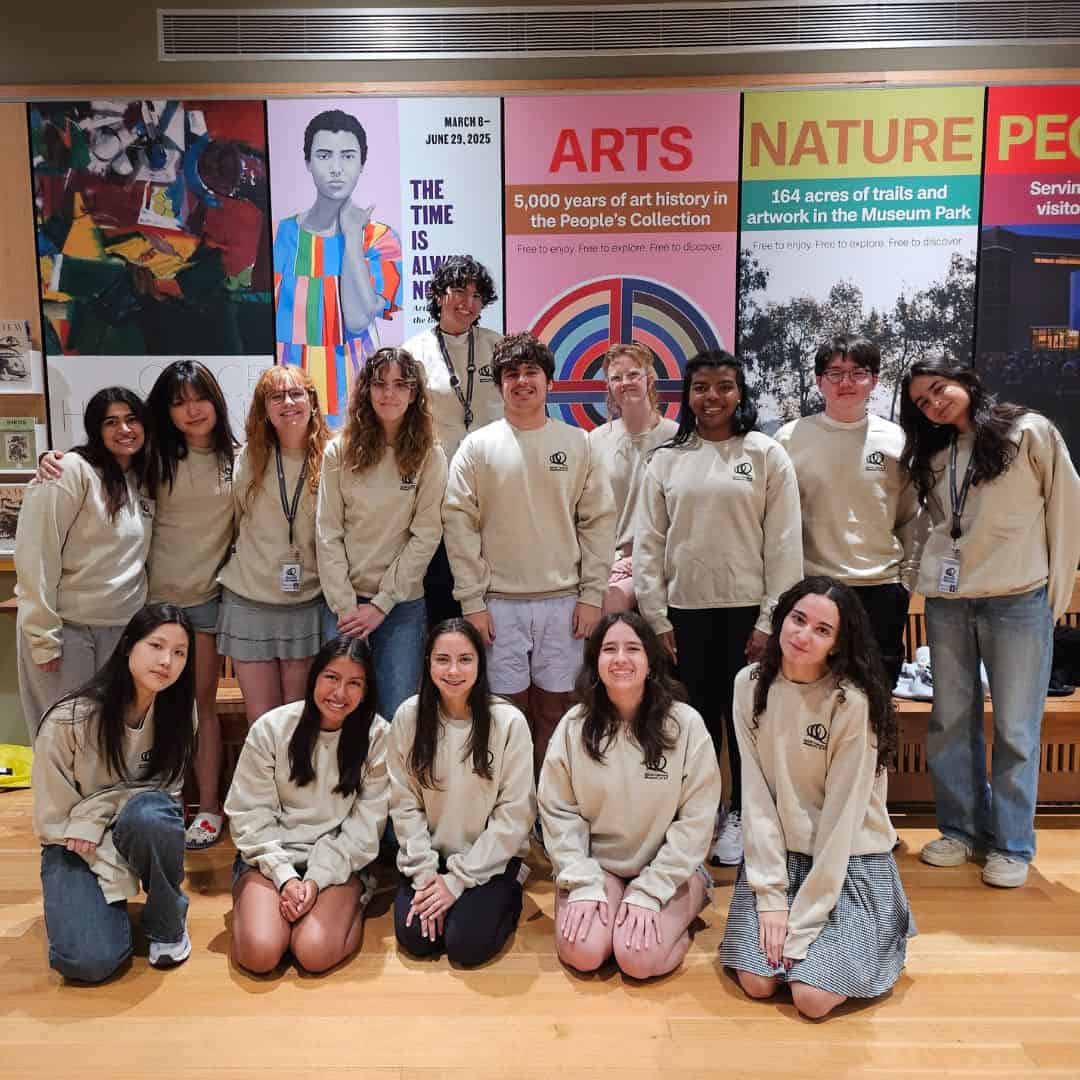 A group of fifteen young people wearing the same beige sweatshirt pose together.