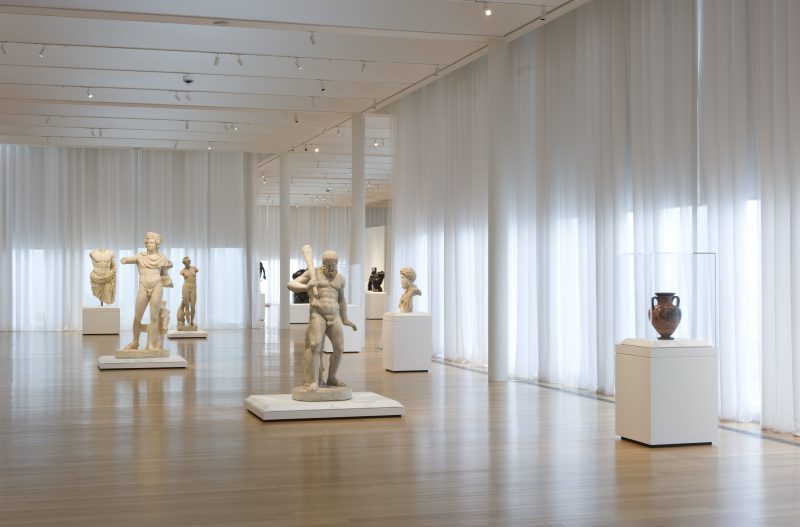 An art gallery containing classical pottery and white marble sculptures displayed on white pedestals. Large windows covered with sheer curtains line the gallery.