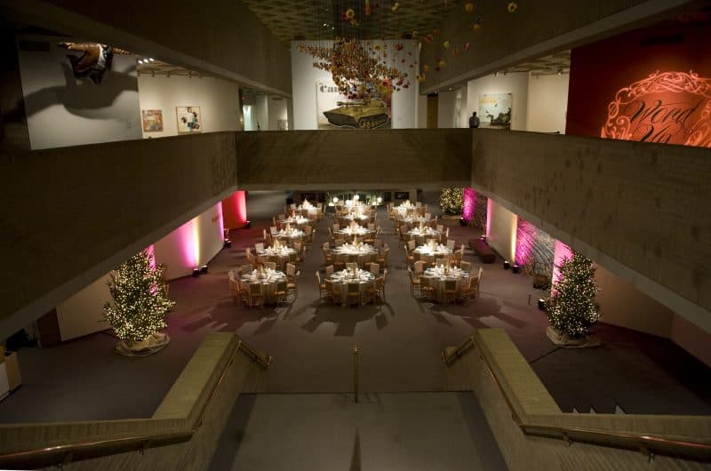 The interior atrium of a three-story building. The bottom floor is decorated with prepared, round tables and Christmas trees. Various works of art are exhibited on the second floor.
