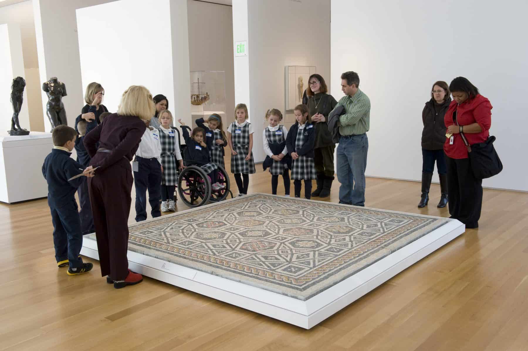 A group of elementary school students and chaperones gather around a large floor mosaic from Classical antiquity that is displayed on the floor of a Museum gallery.