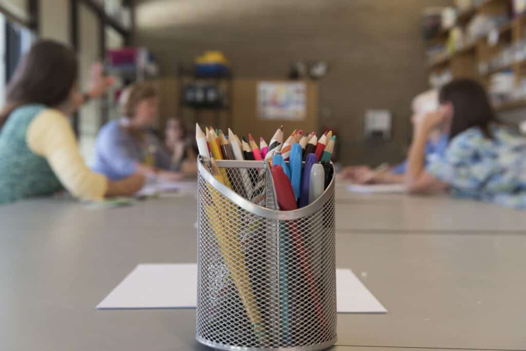 A trisected steel cup constructed with a chain-link pattern holds colored pencils, Sharpies, and pens. In the background there are five visible, yet blurry figures. 