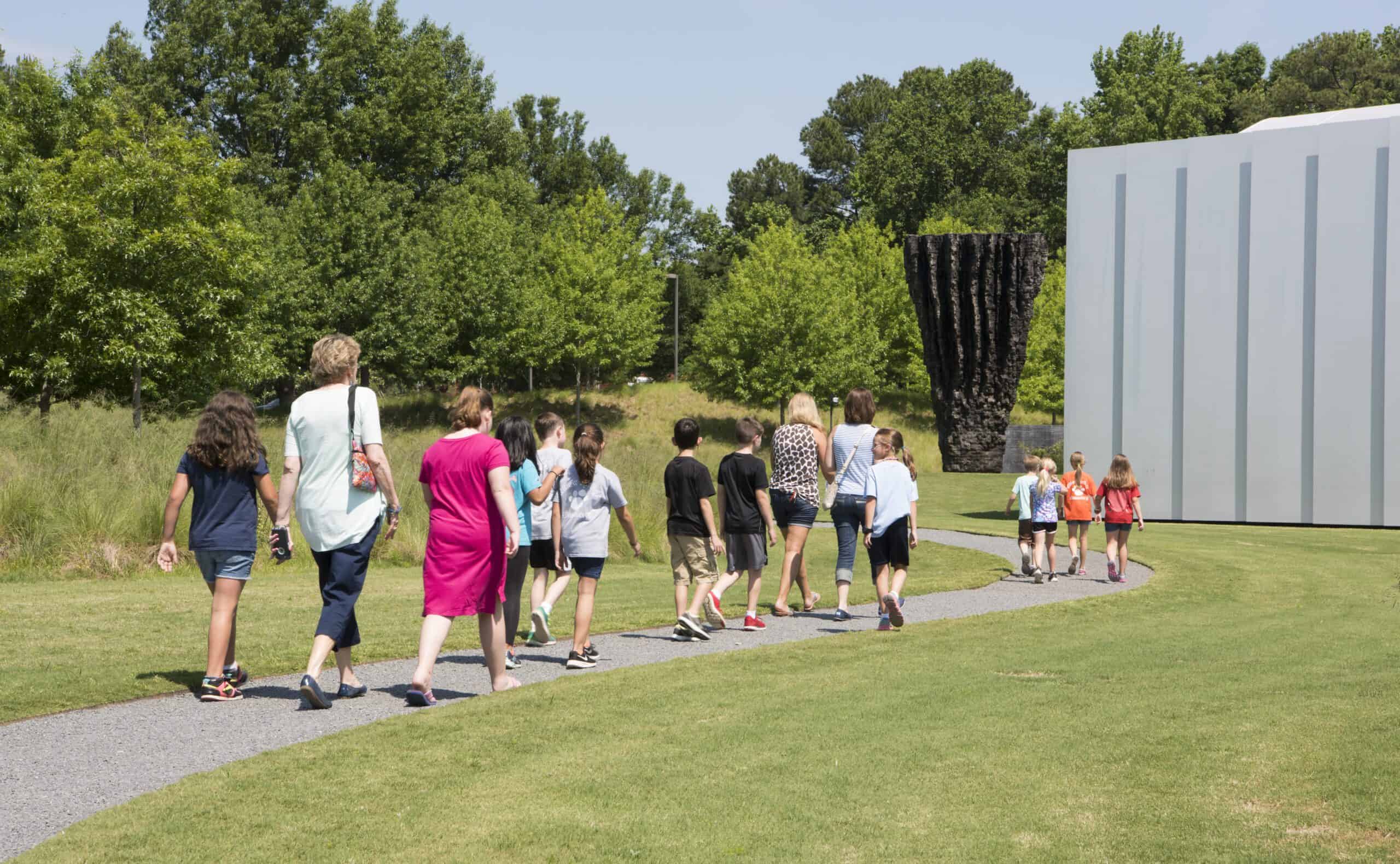 A group of children and three adults walk on a narrow gravel path in the direction of a modernist aluminum facade and a tall, craggy sculpture resembling a hunk of earth.