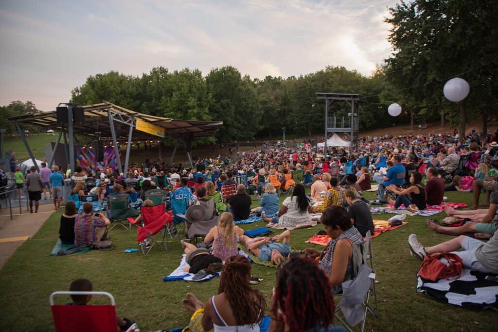 A crowd of people seated on a sprawling outdoor lawn face a covered amphitheater stage. In the background there are trees set against a dusky sky.
