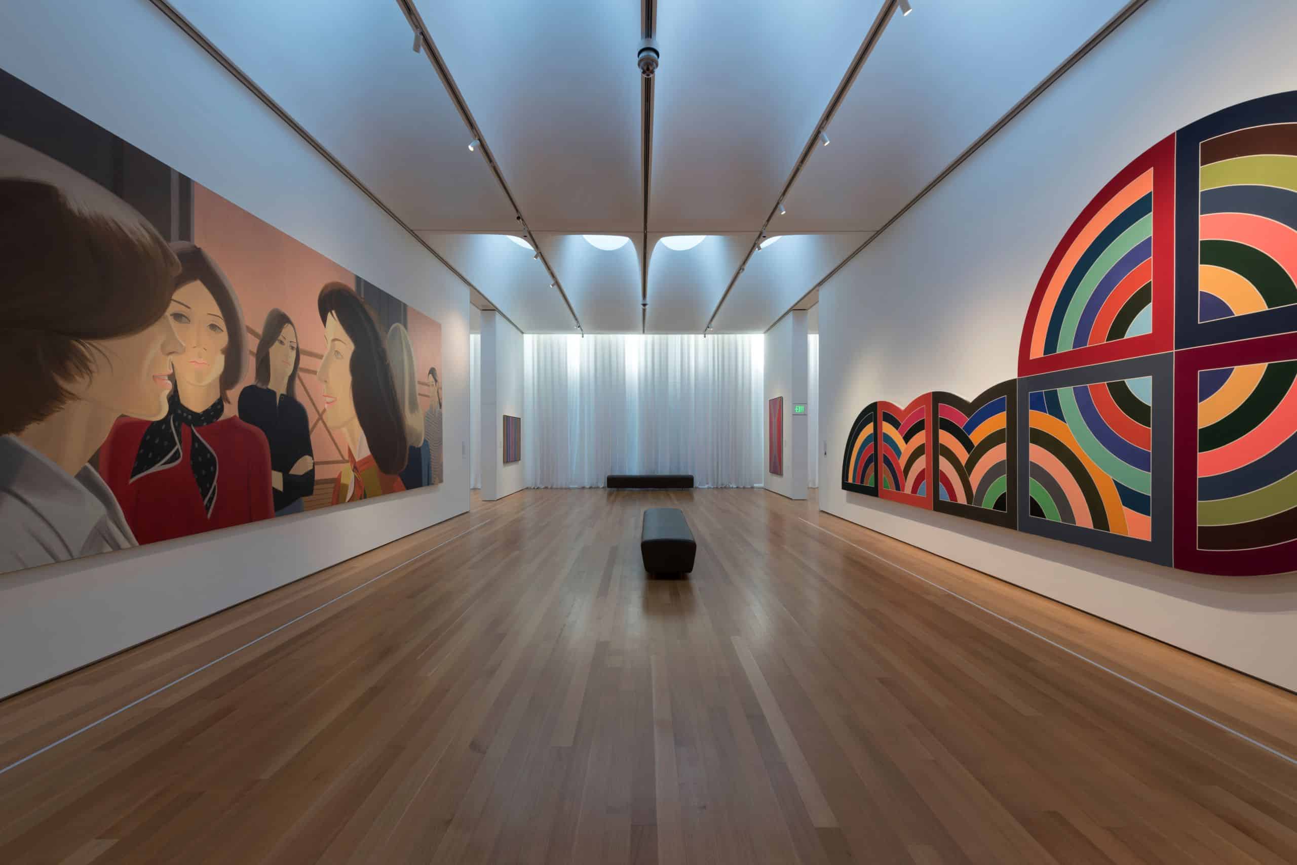 A wood-floored art gallery with various multi-media works on display. A large oil painting of a group of women hangs opposite an abstract painting of overlapping, multicolored half circles along a horizontal line.