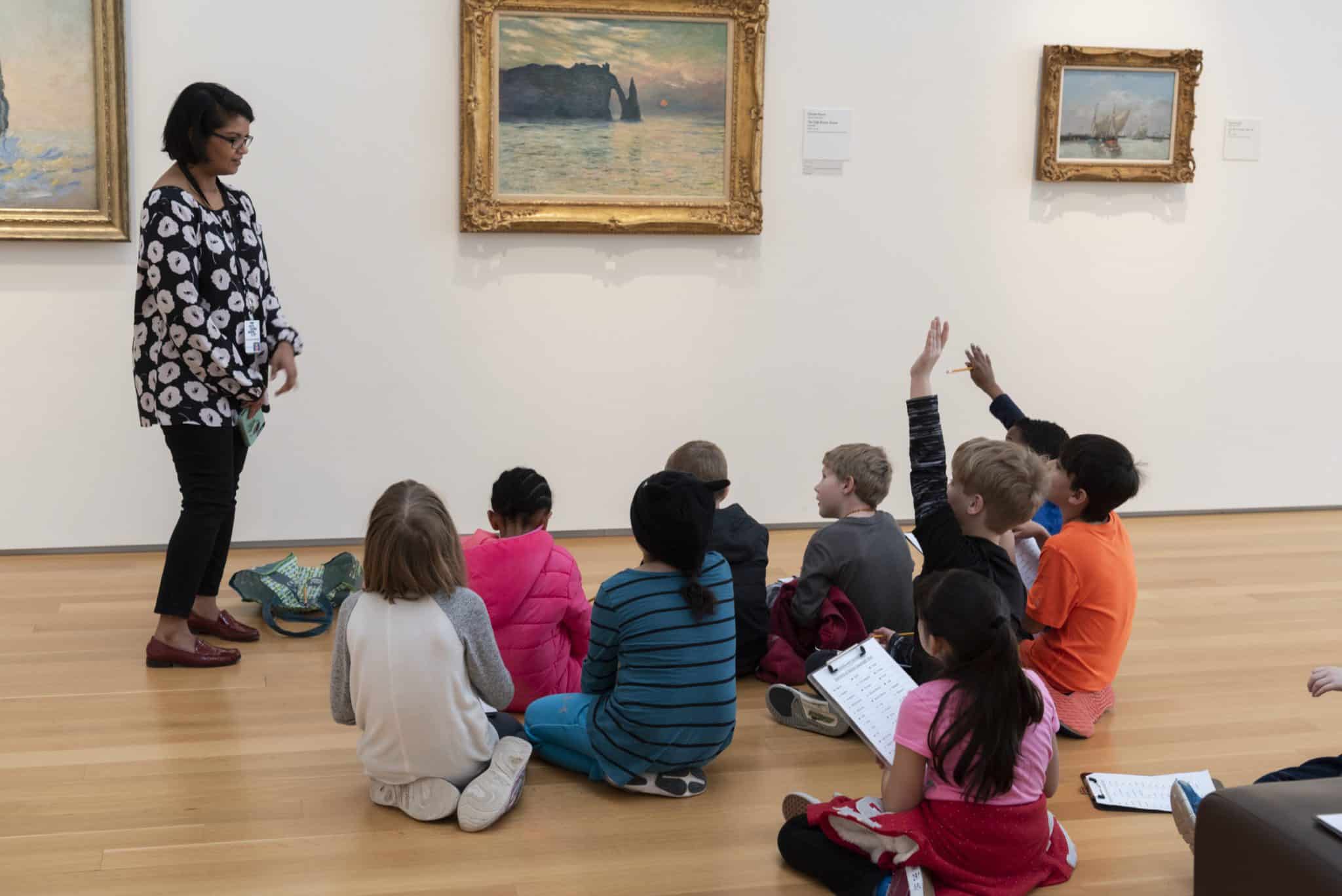 A person standing in front of an Impressionist painting addresses a group of young children seated on the floor of a gallery. Two of the children raise their hands.
