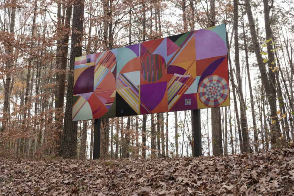 Rectangular billboard with a painted, colorful, geometric pattern displayed on metal poles in a wooded landscape. 
