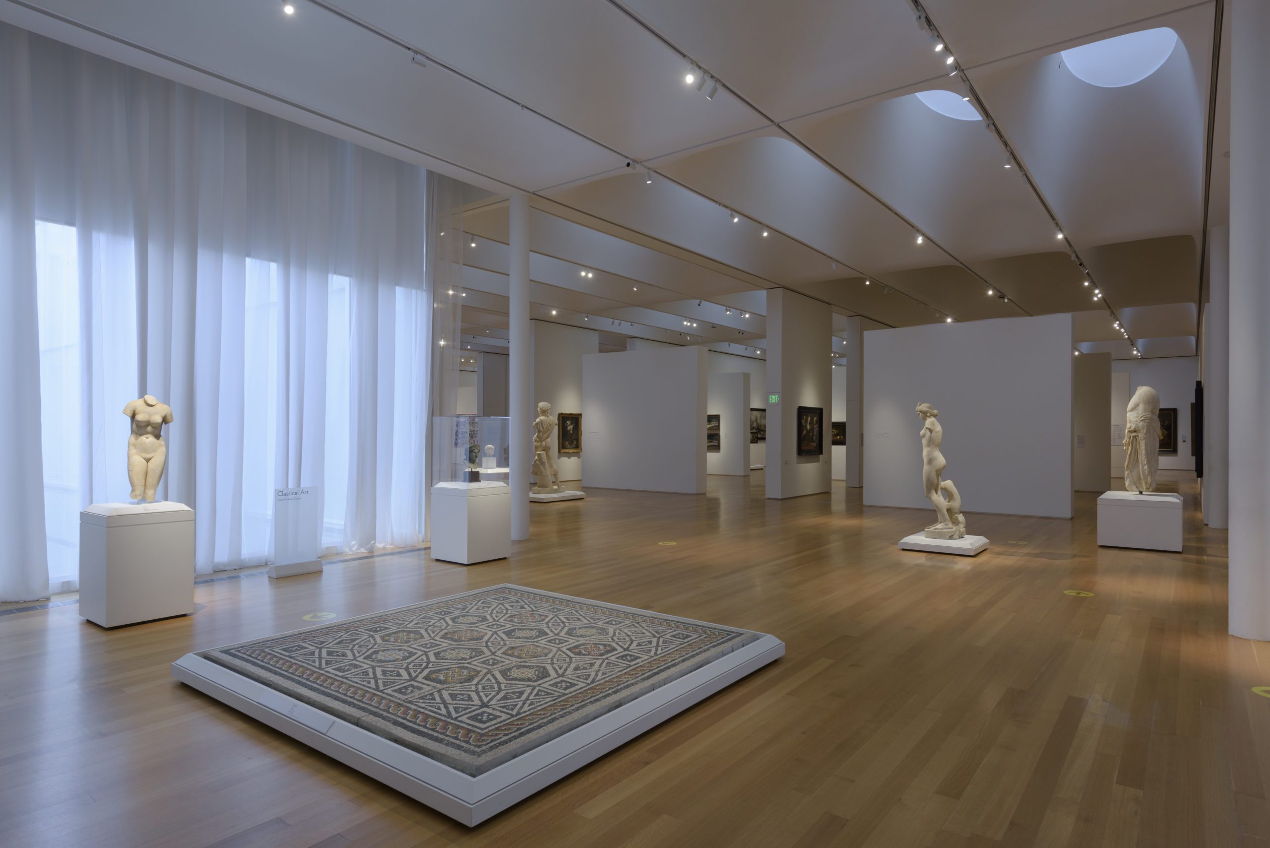 An interior sculpture court populated by various white marble sculptures on pedestals and an ancient mosaic. In the background there are paintings hanging on white walls.