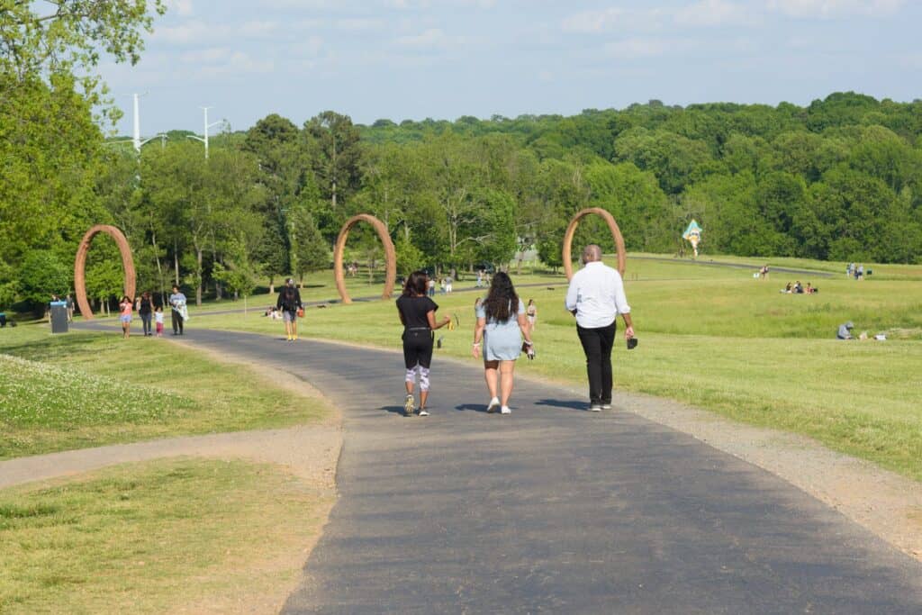 Three people walking on a paved path through rolling fields. In the background there are other people walking along the path, which eventually passed underneath three large, red-orange, ring-shaped sculptures.