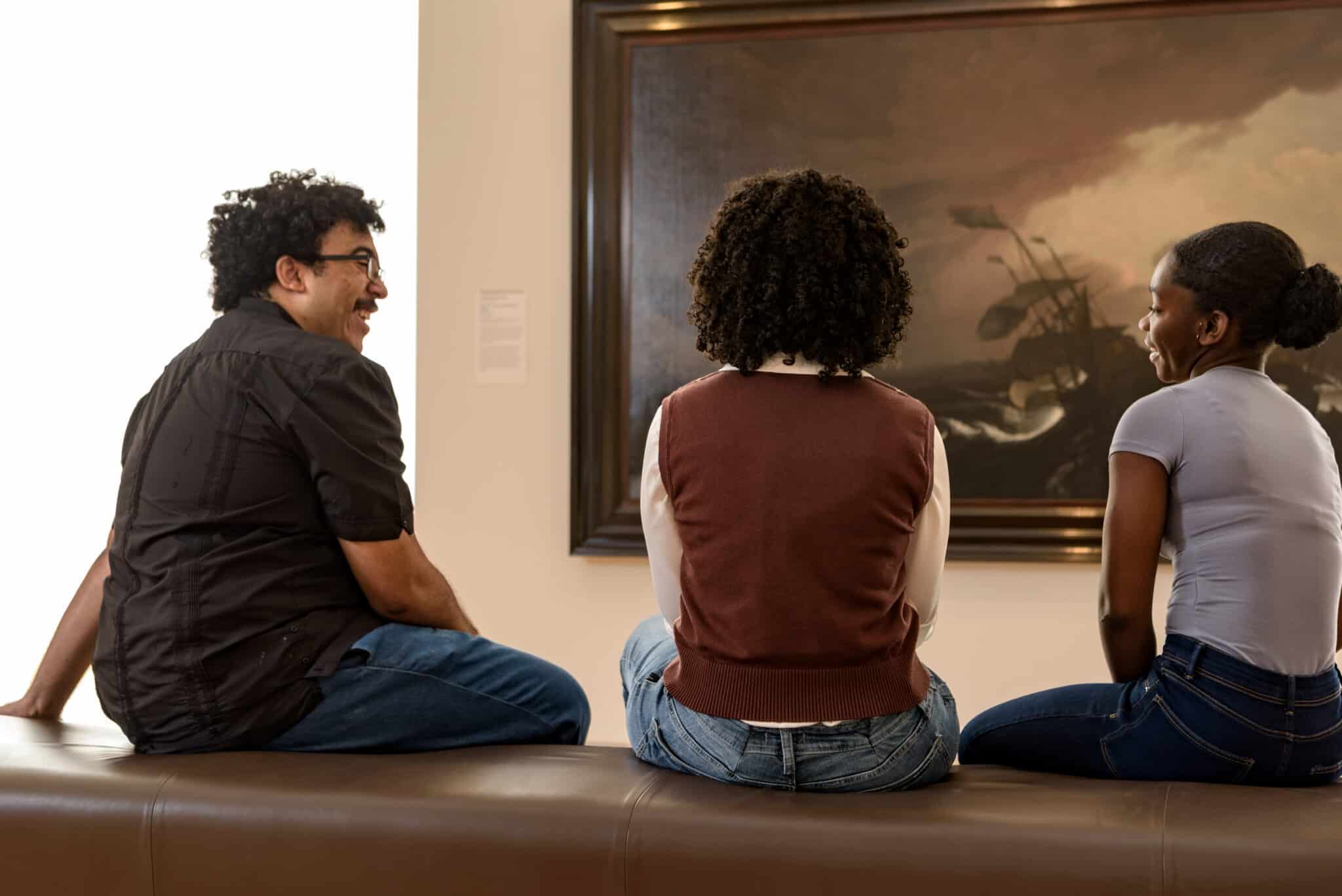 Three people sit on a bench, laughing and talking. On the wall in front of them hangs a large oil painting of a wooden ship in a storm.