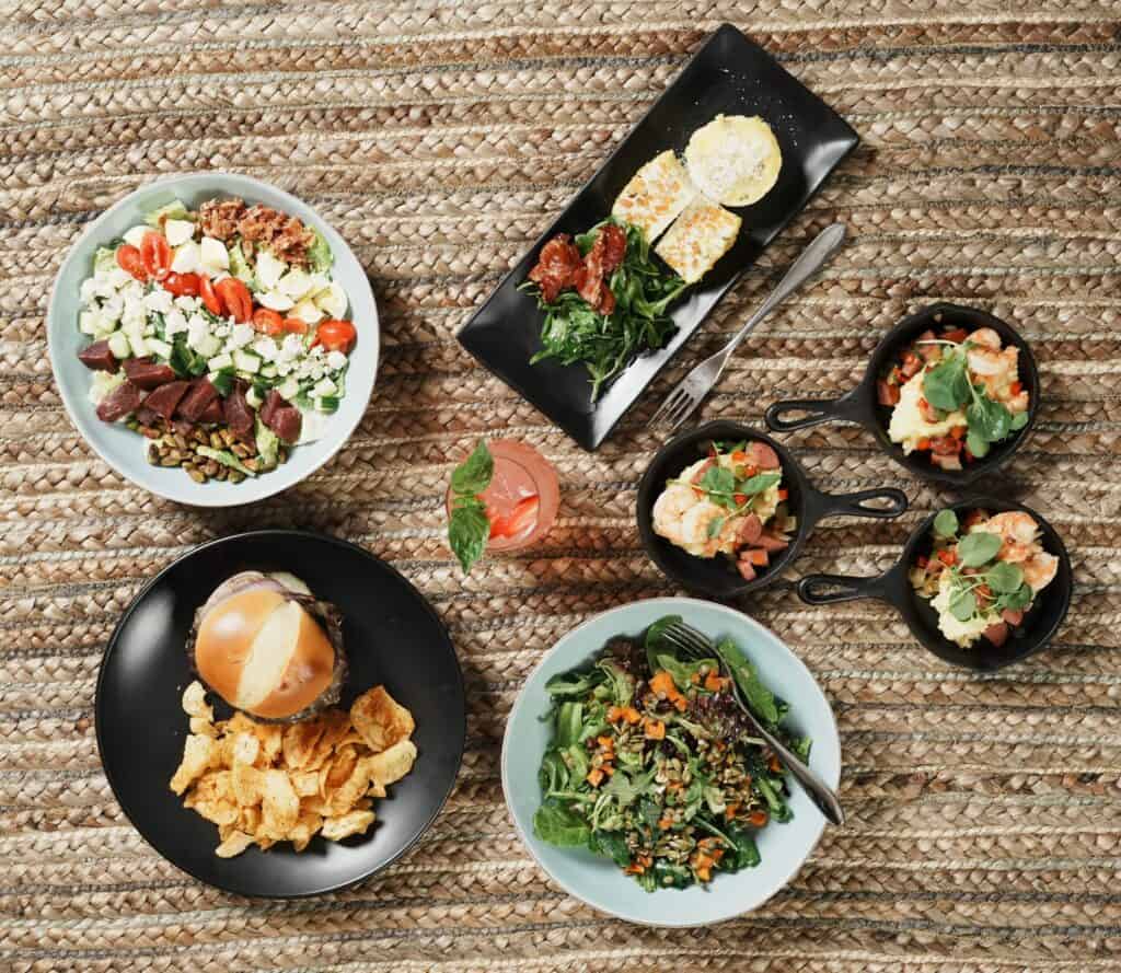 Several plates, bowls and pans of prepared food and one drink neatly arranged on a woven placemat.