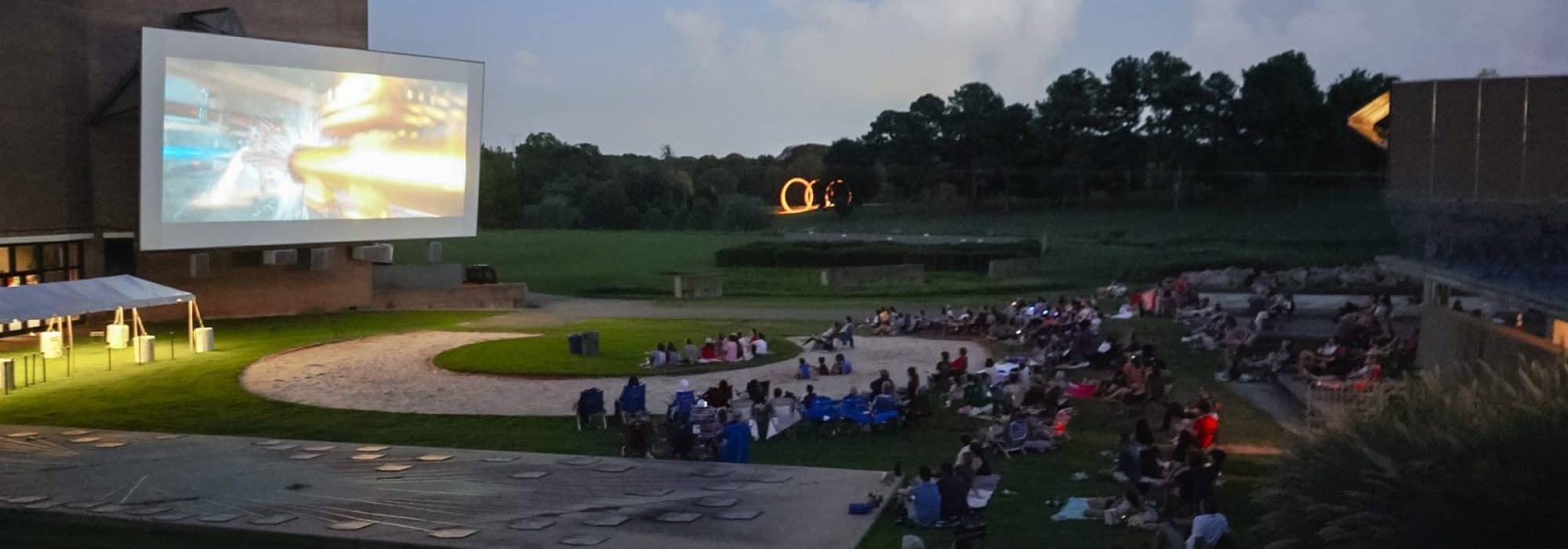 Wide view of a crowd sitting on a lawn and watching a film, which is being projected on a large white screen attached to the side of a two-story brick building.