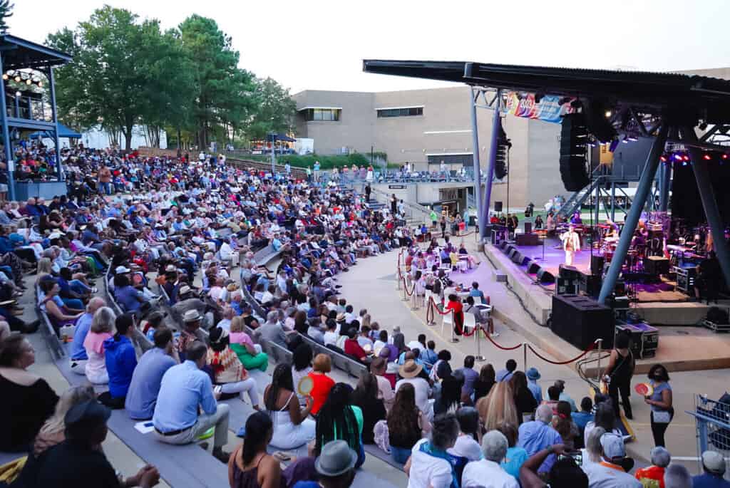 A large group of people sitting in rows of concrete and steel bleachers watch a band perform in an outdoor amphitheater.