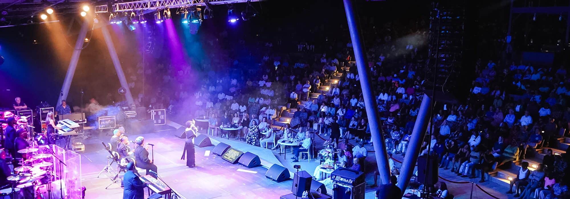 Musicians and singers perform for a crowd seated in an outdoor amphitheater at night.