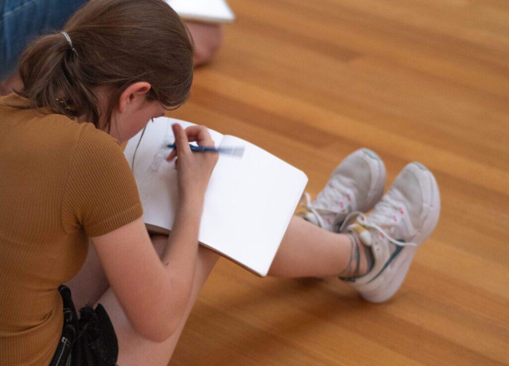 A teen sits on a wooden floor and draws in a notebook that rests on their knees.