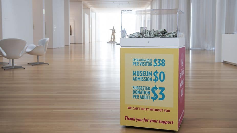 A filled donation box with a graphic decal that suggests a donation amount per museum visitor and thanks museum attendees for their support. In the background there are two white swivel chairs and white marble sculptures.