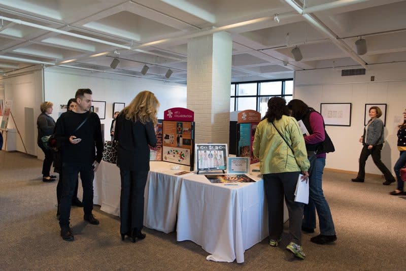Four people stand over tables covered in white tablecloths and inspect colorful tri-fold panels displaying images and text.