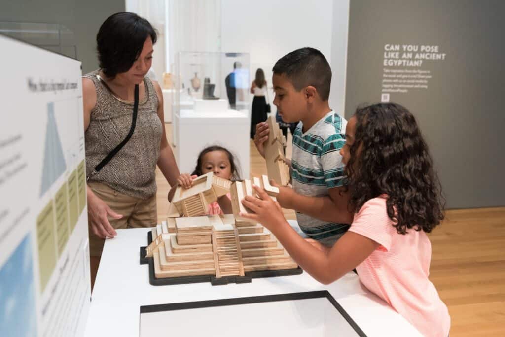 Three children and one adult construct a replica of an ancient American temple made of wood. 